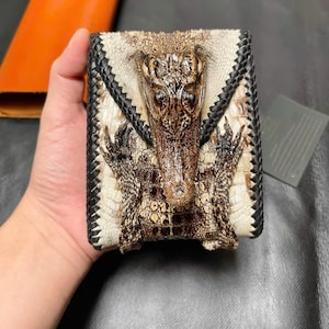 May include: A handcrafted wallet featuring a crocodile design. The wallet has a white and brown color scheme with black braided edges. The crocodile's head and body are prominently displayed on the front of the wallet.