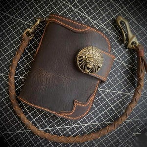 Puede incluir: Una cartera de cuero marrón con un emblema de calavera decorativo y una cadena de cuero trenzado. La cartera tiene costuras naranjas y un cierre metálico. El diseño es robusto y detallado.