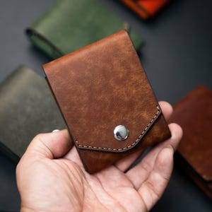 Puede incluir: Una cartera de cuero marrón con cierre de botón a presión plateado. La cartera tiene una superficie texturizada y se sostiene en una mano. Otras carteras verdes y naranjas son visibles en el fondo.