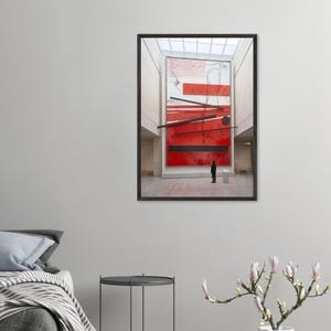 May include: A framed abstract art print featuring a large red and black geometric design. A small figure stands in front of the artwork in a spacious, well-lit gallery setting. The artwork is the focal point.