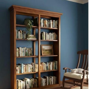 Puede incluir: Una gran estantería de madera llena de libros, colocada contra una pared azul. La estantería tiene varios estantes, una imagen enmarcada y una planta en maceta. Una mecedora con un gato está en la esquina.