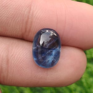 May include: An oval-shaped, cabochon-cut gemstone with a deep blue color and visible internal white veining. The stone has a smooth, polished surface that reflects light, showcasing a translucent quality with varied shades of dark and light blue.