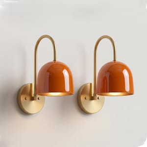 May include: A pair of wall-mounted lamps featuring glossy orange dome-shaped shades and curved brass-toned arms. Each lamp is attached to a circular brass-toned wall plate, showcasing a clean design with a polished finish and warm light.