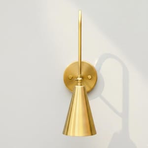May include: A wall-mounted light fixture with a brushed brass finish. It features a circular backplate, a vertical arm extending upward, and a downward-facing conical shade. The design is simple and metallic, showcasing a warm, reflective surface.
