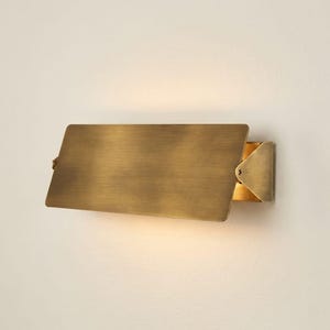 May include: A rectangular wall sconce featuring a brushed brass finish. The fixture has a flat, horizontal metal plate that extends outward from a side-mounted bracket, casting a warm, soft glow from behind the plate onto the wall surface.