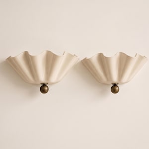 May include: A pair of wall sconces featuring cream-colored, ruffled, bowl-shaped shades with a glossy finish. Each sconce is supported by a central, dark bronze-toned spherical accent piece at the base, creating a symmetrical design.