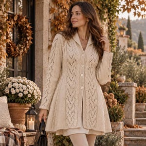 Op de afbeelding: Een beige vest met kabelpatroon, sjaalkraag, lange mouwen en een knoopsluiting aan de voorzijde. Het kledingstuk heeft een verticaal textuurpatroon over het lijfje en de mouwen, met een uitlopende zoom die boven de knieën valt.