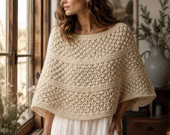 May include: A cream-colored, short-sleeved crochet poncho featuring a wide boat neckline and alternating bands of openwork lace and solid garter stitch patterns, worn over a simple white dress with a tiered skirt design.