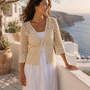 May include: A cream-colored, open-knit crochet cardigan featuring three-quarter length sleeves, a V-neckline, and a single front button closure, worn over a simple white dress. The garment displays a delicate, repeating geometric pattern.