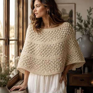 Peut inclure: Un poncho tricoté à la main de couleur crème, présentant un motif texturé à points ajourés avec des bandes horizontales alternées en point mousse. Le vêtement possède une large encolure bateau et une silhouette ample et fluide sur les épaules.