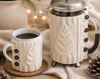 May include: A glass French press and a ceramic mug, both encased in cream-colored knitted cozies featuring a vertical leaf pattern and dark brown buttons. The French press has a silver metal lid and a clear handle, while the mug sits on a wooden coaster.