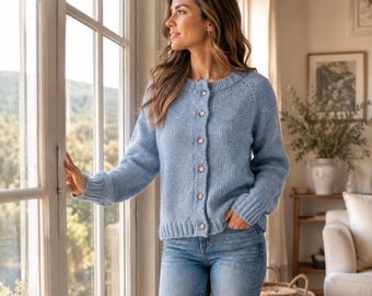 May include: A light blue knit cardigan featuring a round neckline, long sleeves with ribbed cuffs, and a front button closure with five small circular buttons. The garment is paired with blue denim jeans, creating a casual ensemble.