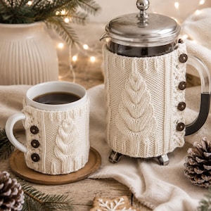 Peut inclure: Une tasse en céramique blanche et une cafetière à piston en verre, toutes deux recouvertes de housses en tricot crème avec un motif de feuilles verticales et des boutons en bois brun foncé. La cafetière a un couvercle argenté.