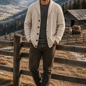 May include: A cream-coloured cable knit cardigan with a shawl collar and dark buttons, worn over a dark grey t-shirt, paired with brown trousers and brown leather boots. The cardigan features a vertical ribbed texture and long sleeves.