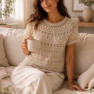 May include: A short-sleeved, cream-colored crochet top featuring a round neckline, intricate openwork patterns, and small raised bobble details across the chest and shoulders. The garment has a straight hem and a textured, hand-crafted appearance.