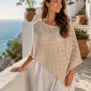 May include: A beige knit poncho featuring an intricate diamond-shaped openwork pattern and a wide, rounded neckline. The garment has a loose, asymmetrical drape that falls over the shoulders and upper body, paired with a plain white skirt.