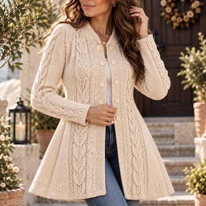 May include: A cream-coloured cable knit cardigan featuring a flared silhouette, long sleeves, and a front button closure. The garment is crafted with a textured vertical cable pattern and a smooth ribbed trim along the neckline and button placket.