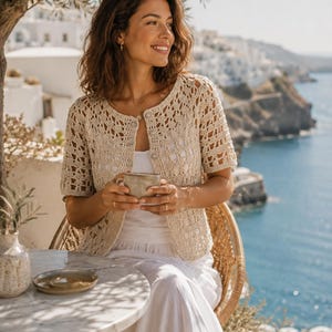 Può includere: Un cardigan beige a maniche corte caratterizzato da un intricato motivo a uncinetto traforato, scollo rotondo e chiusura frontale con bottoni. Il capo è indossato sopra un top bianco semplice, mostrando un design in maglia testurizzata.