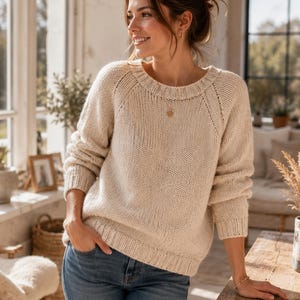 May include: A cream-colored, long-sleeved knit sweater featuring a ribbed crew neckline, ribbed cuffs, and a ribbed hem. The sweater is constructed with a chunky knit texture and raglan sleeves, paired with blue denim jeans.