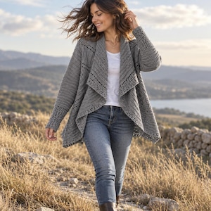 May include: A grey crochet cardigan featuring an open front, long sleeves with textured cuffs, and a draped collar. The garment is worn over a plain white t-shirt and blue denim jeans, showcasing a detailed stitch pattern throughout the fabric.