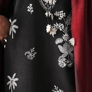 Pode incluir: Túnica preta bordada com desenhos florais brancos. A túnica apresenta um decote detalhado e padrões florais delicados. Um xale bordô é drapeado sobre o ombro. O design é intrincado e elegante.