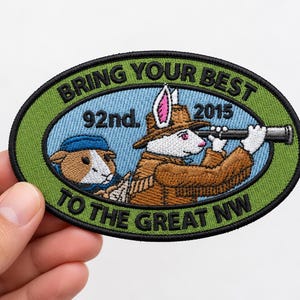 May include: An oval embroidered patch with a green border and light blue centre. It features a white rabbit in a brown coat and hat looking through a telescope, and a brown guinea pig in a blue hat. Text reads: BRING YOUR BEST 92nd. 2015 TO THE GREAT NW.