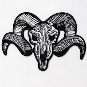 Puede incluir: Un parche bordado que presenta un diseño de cráneo de carnero estilizado en tonos negro, blanco y gris. El diseño muestra texturas complejas similares a huesos y cuernos curvos, con un borde negro grueso que perfila la forma del cráneo.