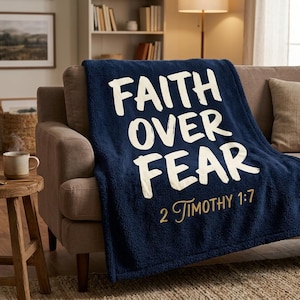 Pode incluir: Um cobertor azul marinho com as palavras "FAITH OVER FEAR" em letras brancas grandes. O texto "2 Timothy 1:7" está em letras douradas menores. O cobertor está sobre um sofá com almofadas.
