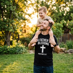 Può includere: Una t-shirt nera a maniche corte con testo bianco che recita A FATHER'S BLESSING e PROVERBS 20:7, centrato sopra e sotto una croce bianca. La maglietta è indossata con jeans blu scuro e un orologio da polso nero sul polso sinistro.