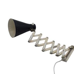 May include: A black and white accordion-style wall lamp. The lamp features a black conical shade with a white interior and a series of white, articulated arms. A white power cord extends from the base.