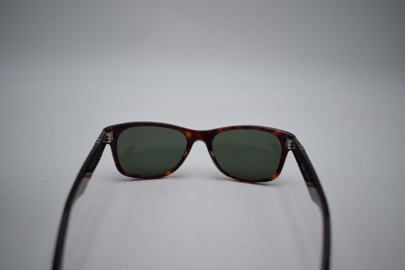 Pode incluir: &Oacute;culos de sol com um design cl&aacute;ssico. As arma&ccedil;&otilde;es apresentam um padr&atilde;o tartaruga, com lentes verdes escuras. As hastes s&atilde;o de cor preta lisa. Os &oacute;culos de sol est&atilde;o sobre um fundo branco liso.