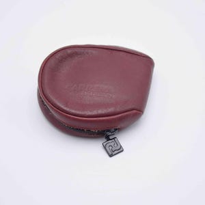 May include: A burgundy leather coin purse with a zipper closure and a black metal zipper pull. The purse has a rounded shape and a small logo on the zipper pull.