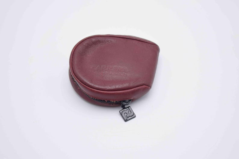 May include: A burgundy leather coin purse with a zipper closure and a black metal zipper pull. The purse has a rounded shape and a small logo on the zipper pull.