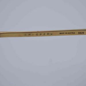 May include: Close-up of a gold-colored eyeglass arm with the text "MADE IN AUSTRIA 5628 41". The arm has a rectangular shape and is attached to a dark brown hinge.