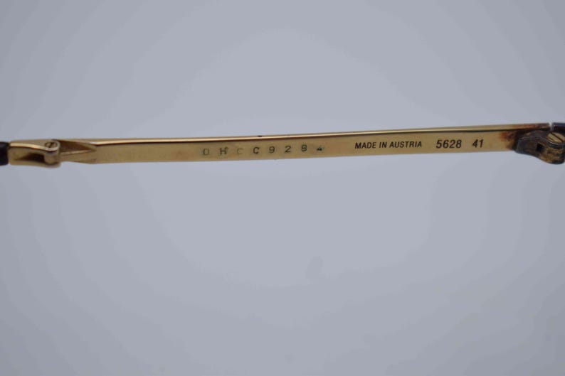 May include: Close-up of a gold-colored eyeglass arm with the text "MADE IN AUSTRIA 5628 41". The arm has a rectangular shape and is attached to a dark brown hinge.