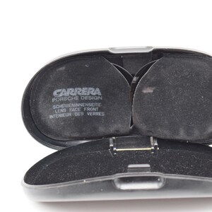 K&ouml;nnte beinhalten: Ge&ouml;ffnete schwarze Carrera Porsche Design Brillenetui. Der Innenraum ist mit schwarzem Stoff ausgekleidet und verf&uuml;gt &uuml;ber zwei kreisf&ouml;rmige F&auml;cher. Das Etui ist klappbar und das Carrera Porsche Design Logo ist sichtbar.
