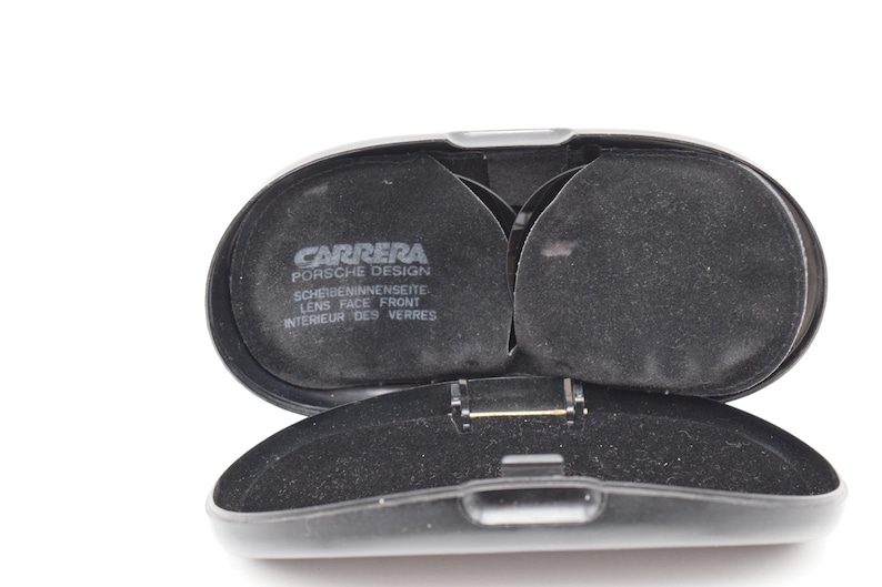 K&ouml;nnte beinhalten: Ge&ouml;ffnete schwarze Carrera Porsche Design Brillenetui. Der Innenraum ist mit schwarzem Stoff ausgekleidet und verf&uuml;gt &uuml;ber zwei kreisf&ouml;rmige F&auml;cher. Das Etui ist klappbar und das Carrera Porsche Design Logo ist sichtbar.