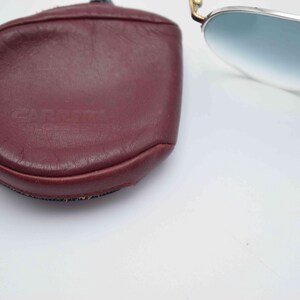May include: A pair of aviator sunglasses with a burgundy leather case. The sunglasses have a silver frame and gradient blue lenses. The case is oval-shaped with the brand name "CARRERA" embossed on the front.