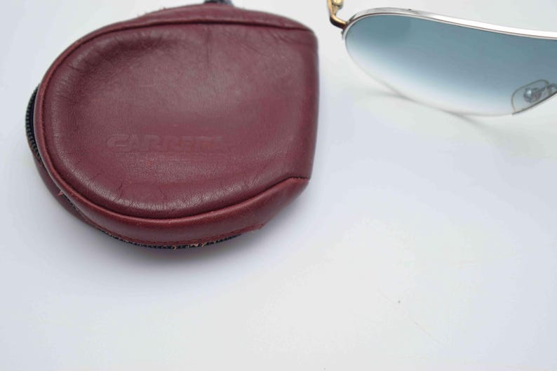 May include: A pair of aviator sunglasses with a burgundy leather case. The sunglasses have a silver frame and gradient blue lenses. The case is oval-shaped with the brand name "CARRERA" embossed on the front.