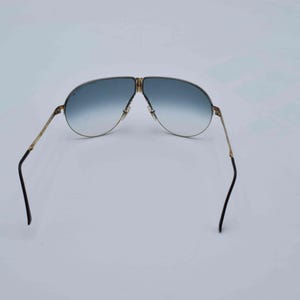 May include: Gold-framed aviator sunglasses with gradient blue lenses. The sunglasses have a double bridge and black earpieces. The lenses transition from a darker blue at the top to a lighter blue at the bottom.