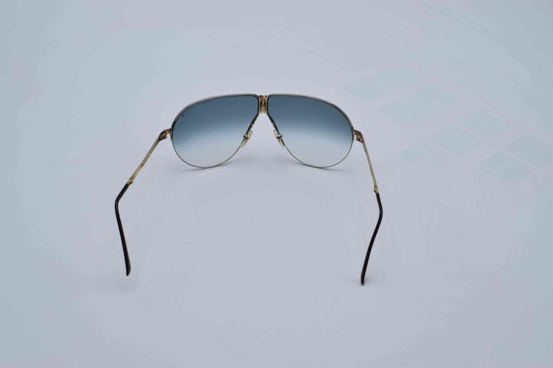 May include: Gold-framed aviator sunglasses with gradient blue lenses. The sunglasses have a double bridge and black earpieces. The lenses transition from a darker blue at the top to a lighter blue at the bottom.