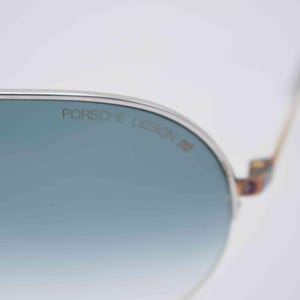 May include: Close-up of Porsche Design sunglasses with a silver frame and blue-tinted lenses. The brand name is printed on the lens. The temples have a patterned design.