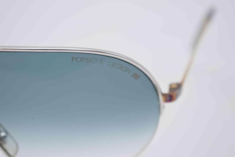 May include: Close-up of Porsche Design sunglasses with a silver frame and blue-tinted lenses. The brand name is printed on the lens. The temples have a patterned design.