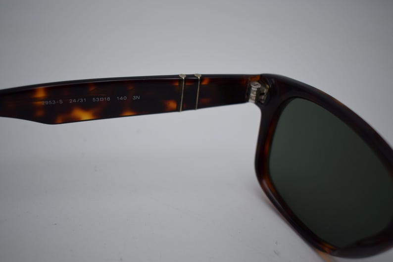Pode incluir: &Oacute;culos de sol tartaruga com lentes escuras. A arma&ccedil;&atilde;o apresenta um design cl&aacute;ssico com uma dobradi&ccedil;a vis&iacute;vel e o texto "2953-S 24/31 53018 140 3N" impresso na haste. As lentes s&atilde;o de cor verde escuro.