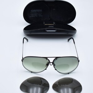 K&ouml;nnte beinhalten: Schwarze Aviator-Sonnenbrille mit gr&uuml;n get&ouml;nten Gl&auml;sern und passendem schwarzem Etui. Das Etui ist ge&ouml;ffnet und gibt den Blick ins Innere frei. Zwei zus&auml;tzliche Gl&auml;ser sind ebenfalls enthalten. Die Sonnenbrille hat einen Metallrahmen.