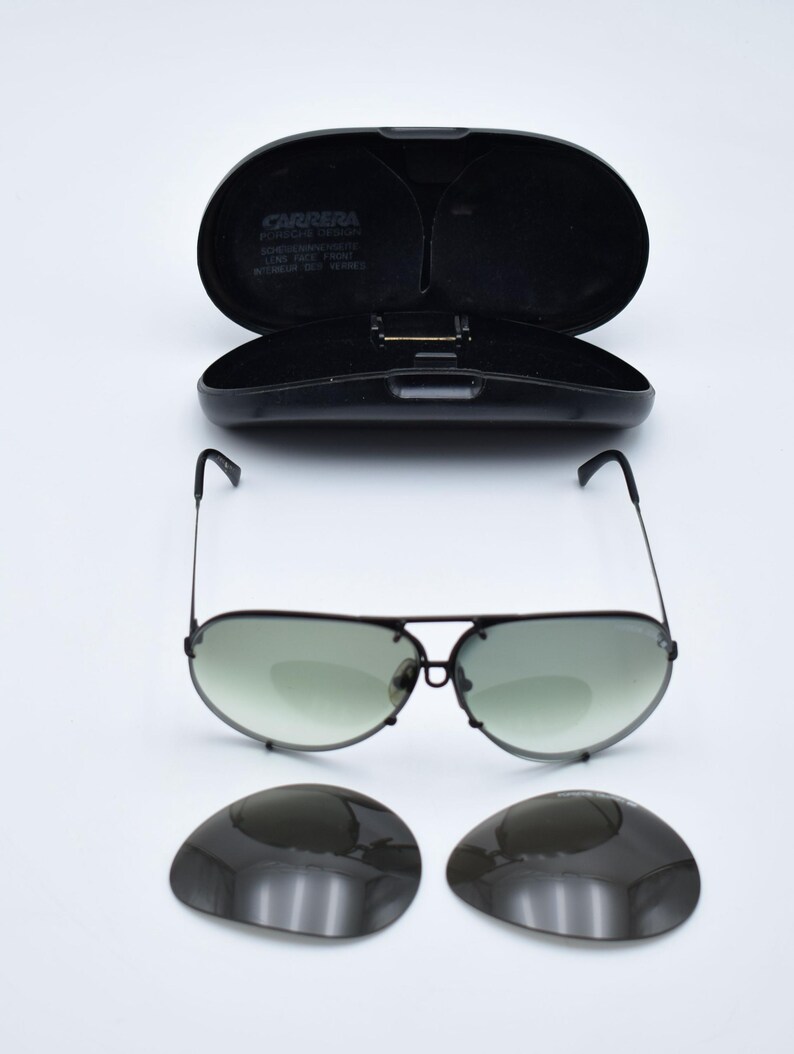 K&ouml;nnte beinhalten: Schwarze Aviator-Sonnenbrille mit gr&uuml;n get&ouml;nten Gl&auml;sern und passendem schwarzem Etui. Das Etui ist ge&ouml;ffnet und gibt den Blick ins Innere frei. Zwei zus&auml;tzliche Gl&auml;ser sind ebenfalls enthalten. Die Sonnenbrille hat einen Metallrahmen.