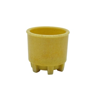 May include: A small, yellow-tan cup with a rounded rim and a textured surface. The cup has a cylindrical body and is supported by three small, rectangular feet. The interior of the cup is visible and empty.