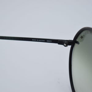 K&ouml;nnte beinhalten: Nahaufnahme einer schwarzen Sonnenbrille mit runden Gl&auml;sern. Der Rahmen ist schwarz mit dem Text "MADE IN AUSTRIA 5621" auf dem B&uuml;gel. Die Gl&auml;ser sind hellgr&uuml;n.