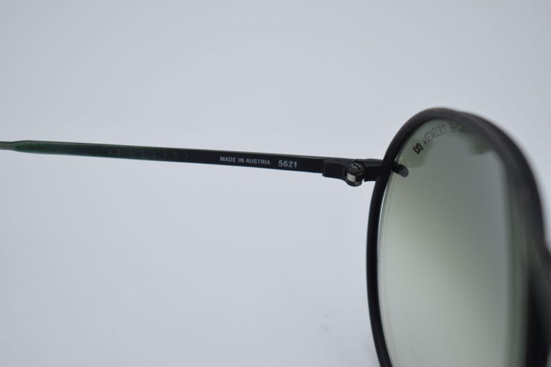 K&ouml;nnte beinhalten: Nahaufnahme einer schwarzen Sonnenbrille mit runden Gl&auml;sern. Der Rahmen ist schwarz mit dem Text "MADE IN AUSTRIA 5621" auf dem B&uuml;gel. Die Gl&auml;ser sind hellgr&uuml;n.