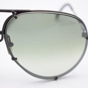 K&ouml;nnte beinhalten: Nahaufnahme einer Vintage-Sonnenbrille von Porsche Design. Die Sonnenbrille hat einen schwarzen Metallrahmen und Verlaufsgl&auml;ser. Der Text "PORSCHE DESIGN" ist auf dem rechten Glas zu sehen. Das Gesamtdesign ist elegant und retro.
